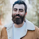 Bearded man with dark hair wearing a tan jacket with white shearling collar, smiling outdoors.