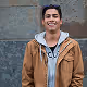 Smiling young man wearing a brown jacket over a gray hoodie standing outdoors.