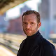 Man with short brown hair and beard wearing a black turtleneck, standing outdoors with blurred buildings in the background.