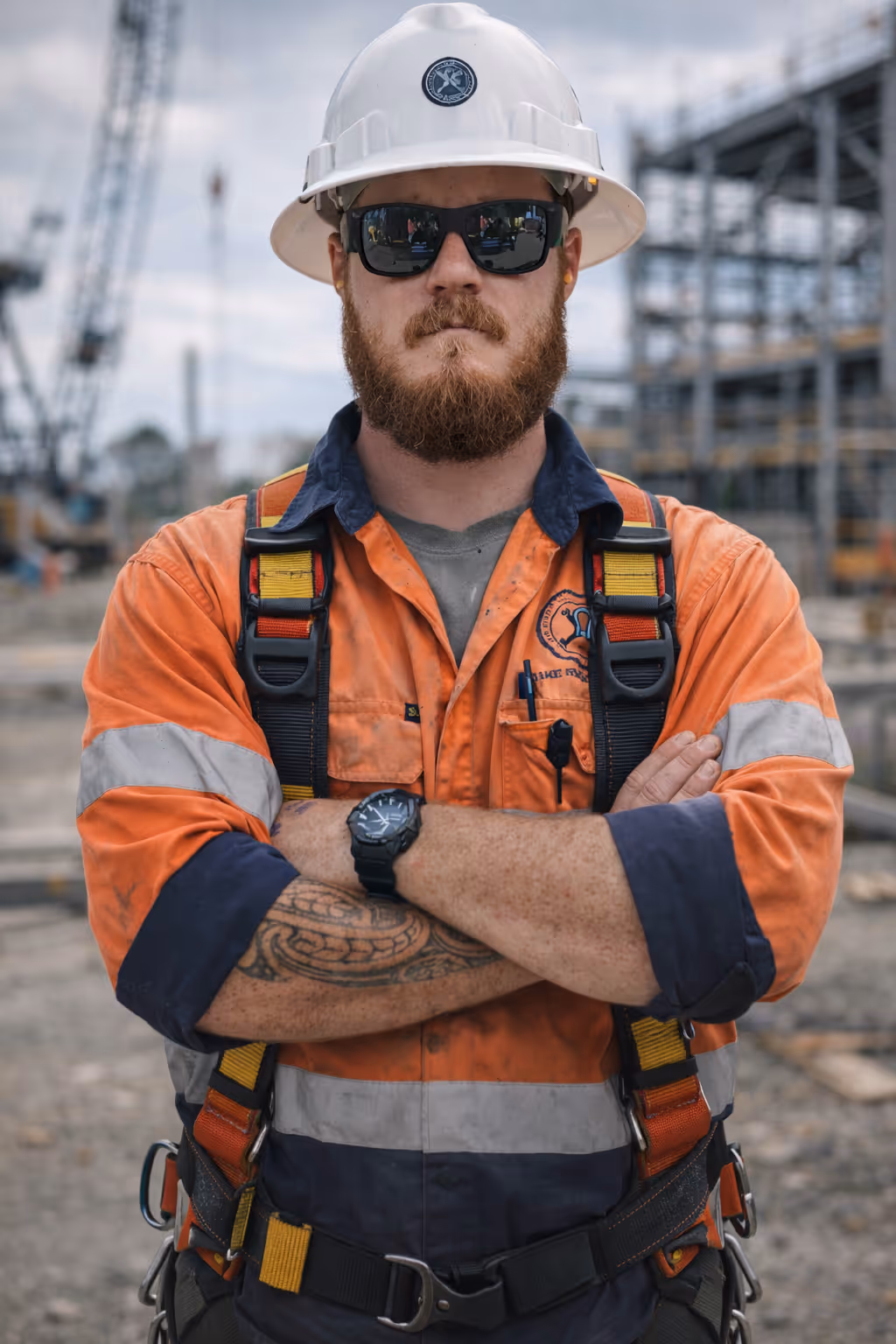 Construction worker with a beard wearing sunglasses, a white helmet, and an orange safety vest crossing his arms at a worksite.