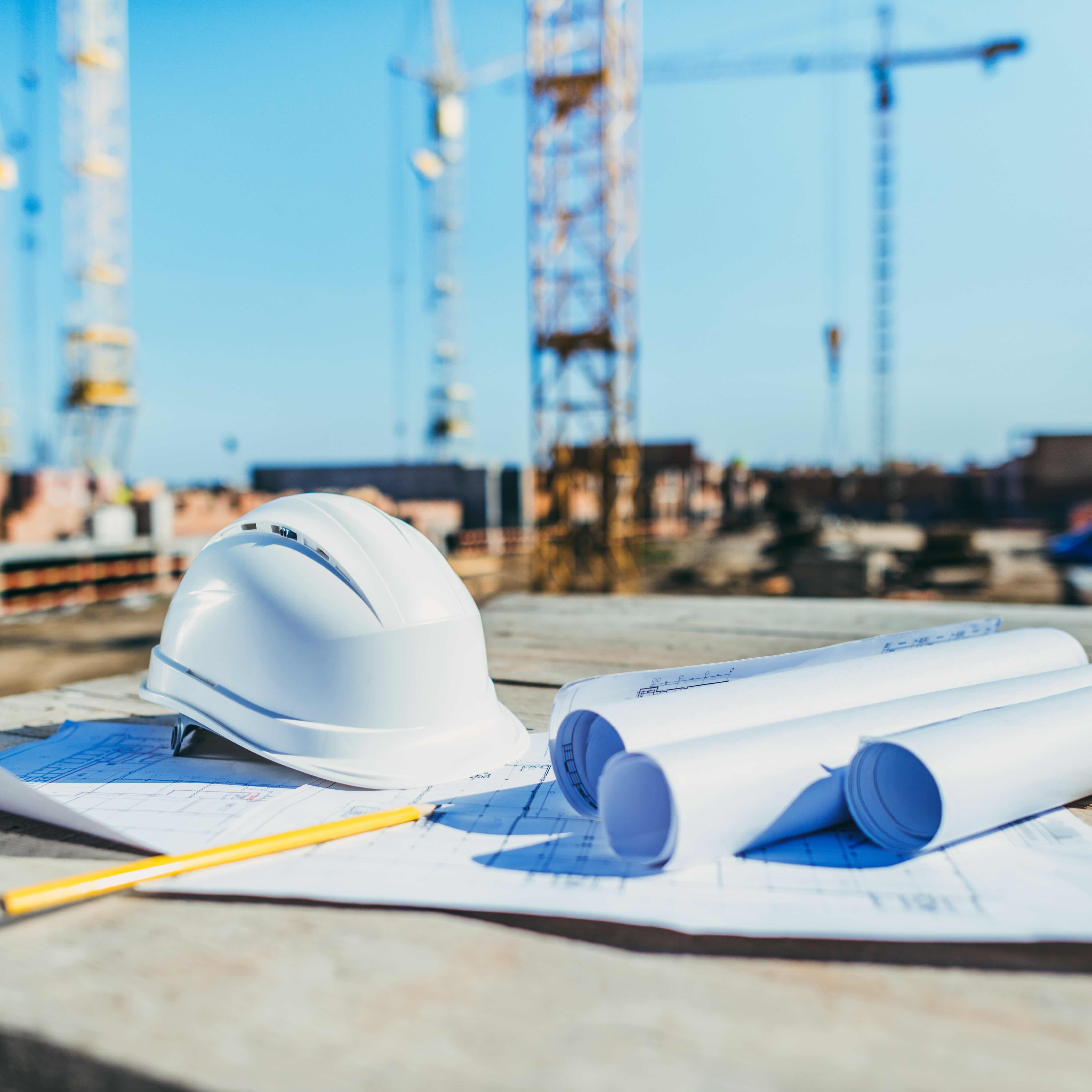 White construction helmet next to rolled blueprints and a pencil on a wooden table at a construction site.